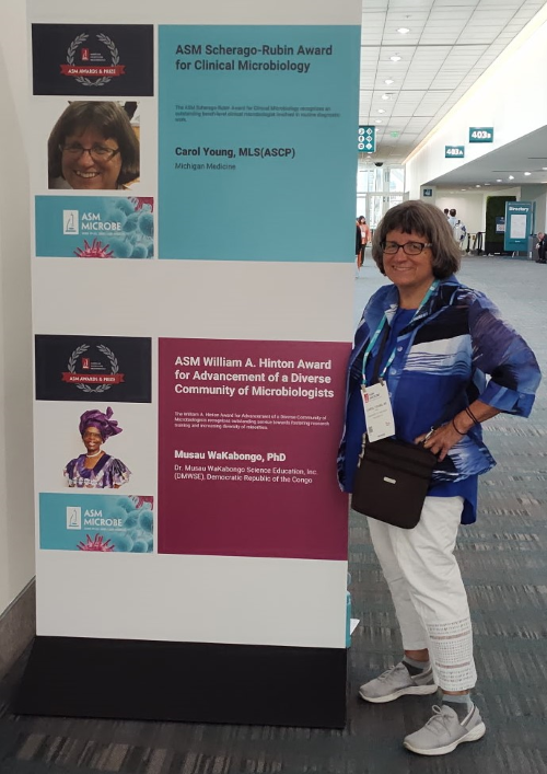 Carol Young stands next to the sign announcing her as the ASM Scherago-Rubin Award for Clinical Microbiology winner.